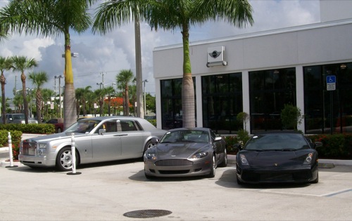 Rolls-Royce Phantom, Aston Martin DB9 és Lamborghini Gallardo egy autókereskedés parkolójában, hülyén beállva